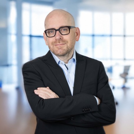 Portrait photo of Moritz Goeb, board member of ISO-Gruppe, responsible for corporate development and finance. He is wearing a black suit, light blue shirt, and black glasses, standing in a modern office environment with his arms crossed.